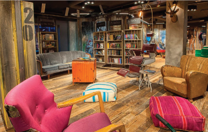 Rustic room with unfinished wood floors, bookshelves, floor cushions, a hot pink arm chiar, an antique barber chair, a large tan arm chair and a grey sofa