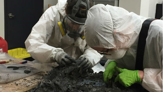 Two professionals in protective clothing work with charred remains
