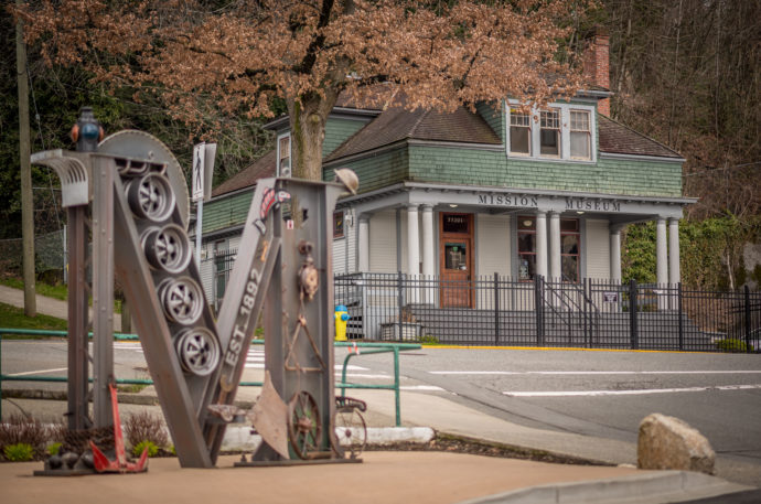 Historic house as seen from the street with trees behind the building. Metallic sculpture of an M on the other side of the street in foreground