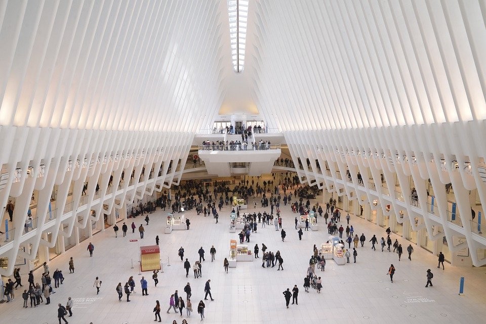 Tiny figures bustling around the entrance of the World Trade Centre, which has open wall slats on either side of the entrance that connect at the ceiling to form an open triangle.