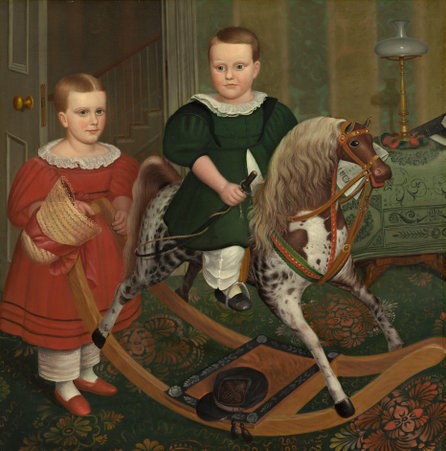 Robert Peckham’s oil painting, The Hobby Horse, depicts two fair-skinned children with short brown hair. The child in the centre of the painting is wearing a white ruffled collar, a dark green dress, and white crimped pantaloons, a type of baggy pant. This child is holding a black horsewhip and rising on a toy rocking horse, with a light brown mane and white body with brown spots. To the left of the child and rocking horse is another child wearing a similar outfit of a white ruffled collar, a dark red dress, and white crimped pantaloons. The child wearing the red dress is holding a straw hat with a red bow on it and is looking directly at the viewer. The two children are inside a late 1800s home, complete with an ornate dark green carpet with red and yellow floral graphics, a brass oil lamp on a wooden table, and blue and yellow striped wallpaper with a gold floral design repeating on every stripe.